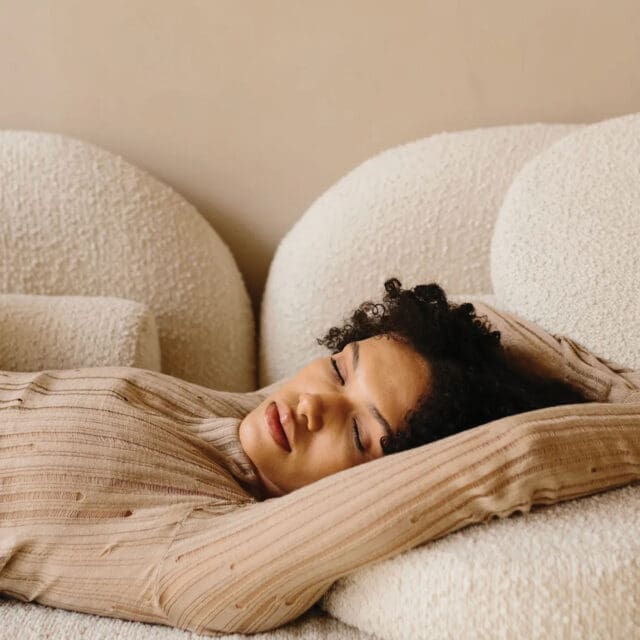 A woman rests on a couch, arms outstretched, peacefully sleeping in a cozy setting.